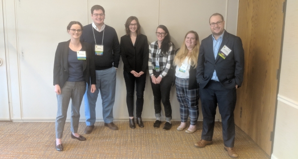 The 2019 GSIC Committee standing in front of a beige wall in conference attire. From left to right, Elizabeth Deacon, Samuel Hahn, Sara Hales-Brittain, E.L. Meszaros, Sarah Keith, and Samuel Kindick.